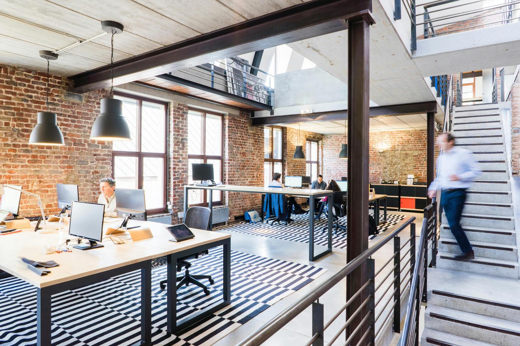 Offenes Büro mit Holz- und Metallmöbeln, großen Fenstern und einem zentralen Treppenhaus. Personen arbeiten am Computer.