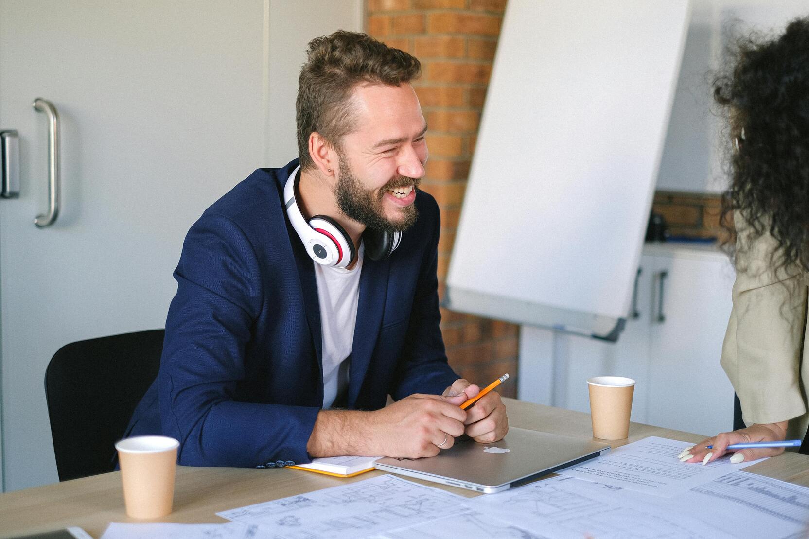 Lachender Mann mit Kopfhörern sitzt an einem Tisch, umgeben von Kaffeebechern und Dokumenten.
